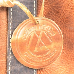 May include: Close-up of a leather product with a circular tag. The tag is embossed with the words "andrew phillips" and "Genuine Leather" around a logo. The leather is a warm brown colour with a textured surface.