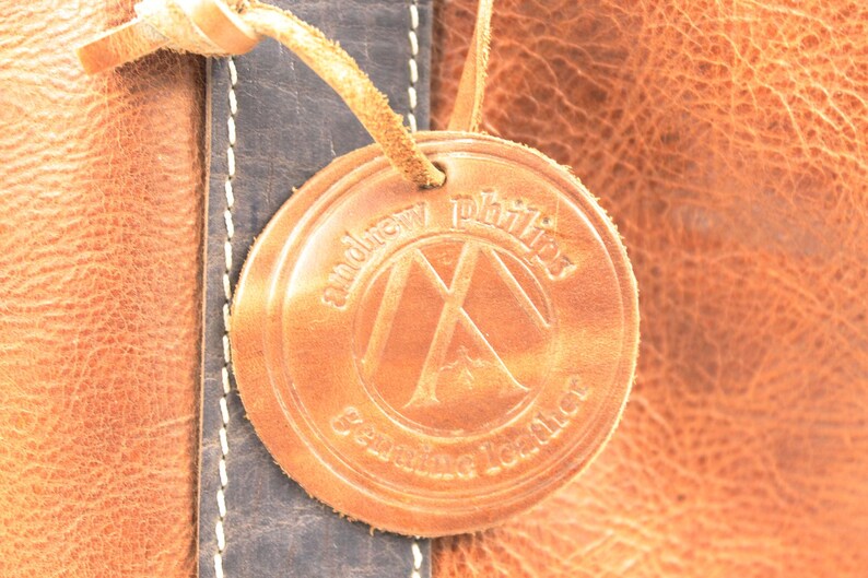 May include: Close-up of a leather product with a circular tag. The tag is embossed with the words "andrew phillips" and "Genuine Leather" around a logo. The leather is a warm brown colour with a textured surface.