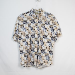 Puede incluir: Una camisa blanca de manga corta con botones y estampado tropical con hojas azules, marrones y amarillas.