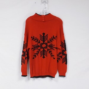 Peut inclure: Pull-over rouge en tricot avec un motif de flocon de neige noir. Le pull a un col montant et une fermeture boutonnée à l'arrière du cou.