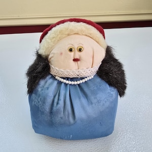 May include: A handmade doll with a round, pale face, large googly eyes, and a small red mouth. It wears a red and white hat, black fur hair, a lace collar, a pearl necklace, and a blue dress. The doll is a decorative item.