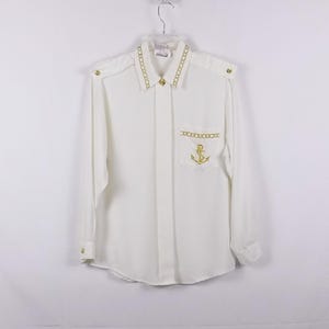 May include: White long-sleeve button-up shirt with a gold chain trim around the collar and a gold anchor embroidered on the pocket.