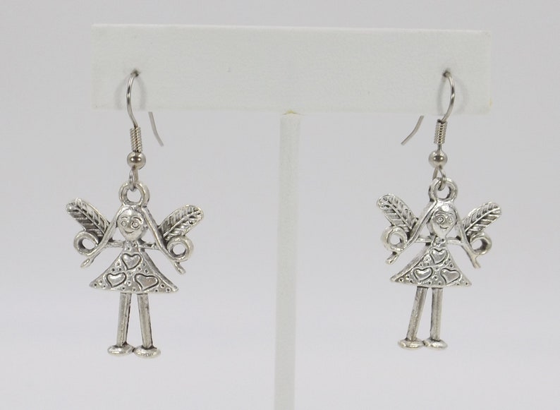 K&ouml;nnte beinhalten: Silberne Ohrh&auml;nger mit einem kleinen, detaillierten Anh&auml;nger einer M&auml;dchenfigur mit Fl&uuml;geln und einem herzf&ouml;rmigen Kleid.