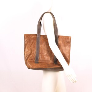 May include: A brown leather tote bag with dark grey straps. The bag has a rectangular shape and a distressed appearance. The straps are attached to the top of the bag and are long enough to be worn over the shoulder.