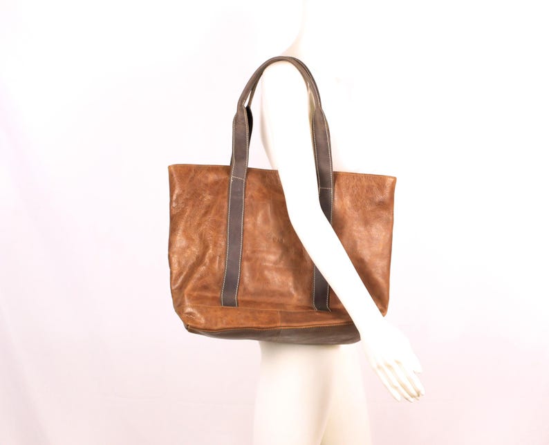 May include: A brown leather tote bag with dark grey straps. The bag has a rectangular shape and a distressed appearance. The straps are attached to the top of the bag and are long enough to be worn over the shoulder.