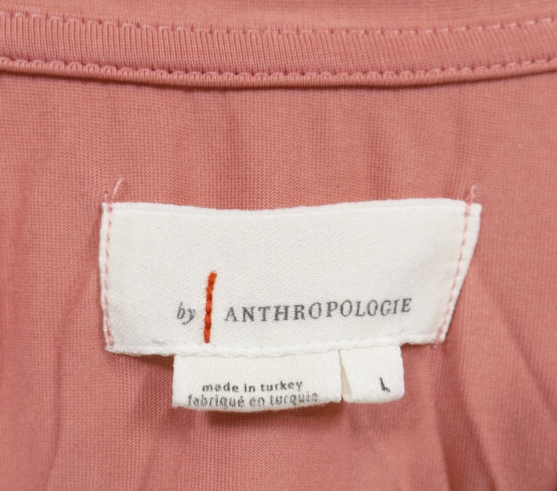 May include: A white tag with the text "by ANTHROPOLOGIE" and "made in turkey / fabriqu&eacute; en turquie". The tag is sewn onto a pink fabric.