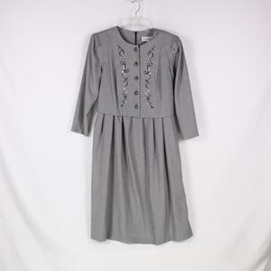 Puede incluir: Un vestido gris con un diseño de bordado floral. El vestido tiene un frente abotonado y una falda fruncida.