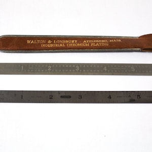 Puede incluir: Una regla de metal de 12,7 cm con una funda de cuero marrón. La funda tiene el texto "WALTON & LONSBURY ATTLEBORO, MASS. INDUSTRIAL CHROMIUM PLATING" impreso en ella.