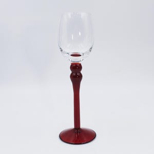 May include: A clear glass wine goblet with a ruby red stem and base. The stem features a bulbous design. The goblet is empty and sits on a white background. The base has a diameter of approximately 3 inches.