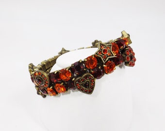 May include: A gold-toned bracelet featuring red and orange crystals and charms. The charms include hearts, stars, and other geometric shapes.