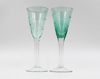 May include: Two clear green glass shot glasses with a swirl design. The glasses have a long stem and a wide base.