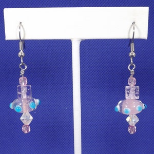 Può includere: Un paio di orecchini pendenti con perline di vetro rosa e blu. Gli orecchini hanno ganci d'argento e sono appesi a un espositore bianco.