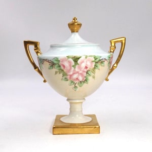 May include: A vintage porcelain urn with a lid, featuring pink rose floral artwork. The urn has gold-coloured handles and a gold base. The lid is topped with a gold finial. The urn is approximately 20 cm tall.