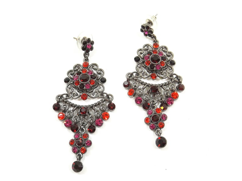 May include: A pair of dangle earrings with a silver tone finish. The earrings feature a large, ornate flower design with red, pink, and black crystals. The earrings are approximately 3 inches long.