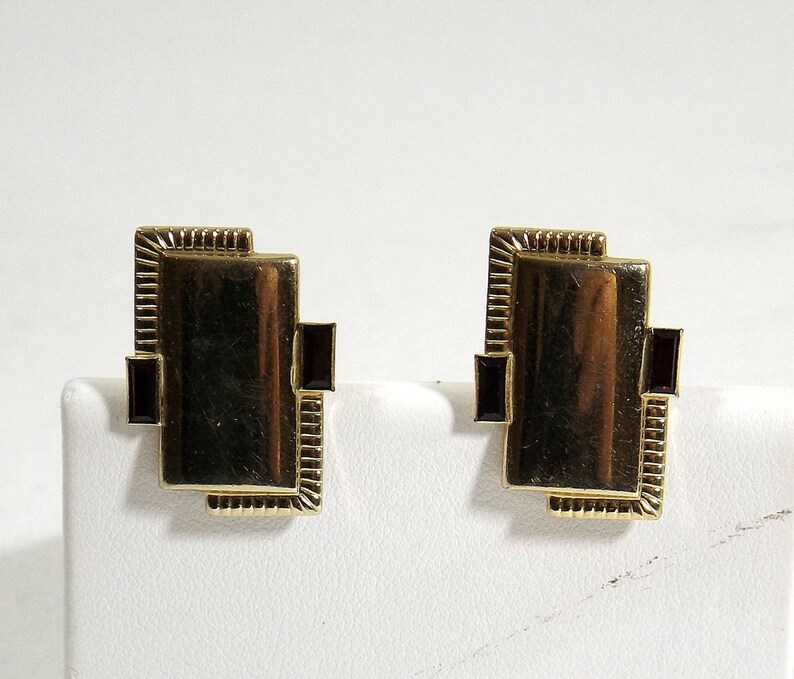 May include: A pair of gold-toned earrings with a rectangular design. Each earring features a dark red stone set in the center.