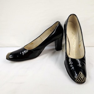 Puede incluir: Par de zapatos de salón negros de charol con detalles de malla dorada en la puntera. Los zapatos tienen un tacón de bloque.