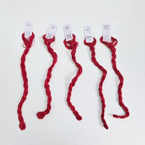 Puede incluir: Cinco hebras de hilo de bordar rojo, cada una con una etiqueta blanca adjunta. Las etiquetas muestran texto y el nombre de la marca "EdMar". El hilo está torcido y dispuesto en un patrón ondulado sobre un fondo blanco.