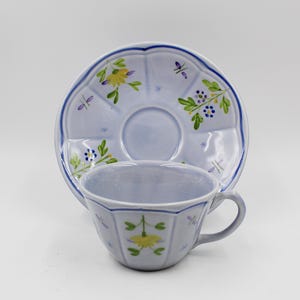 May include: A light blue teacup and saucer set. The cup and saucer have a scalloped edge and are decorated with yellow and green floral designs and small purple accents. The set is made of ceramic.