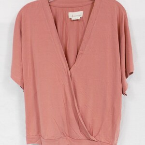 May include: A dusty rose colored, short-sleeved, wrap-style top with a V-neck. The top has a small tag that reads "Anthropologie" on the front.
