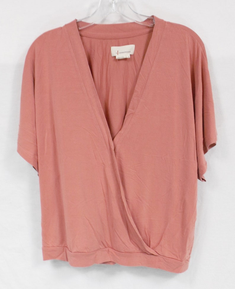 May include: A dusty rose colored, short-sleeved, wrap-style top with a V-neck. The top has a small tag that reads "Anthropologie" on the front.
