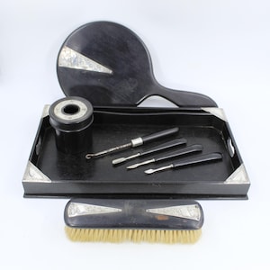 May include: A vintage vanity set with a black tray, hand mirror, and brush. The tray holds a small container and several manicure tools. Silver accents adorn the tray corners, mirror, and brush. The brush has light-colored bristles.