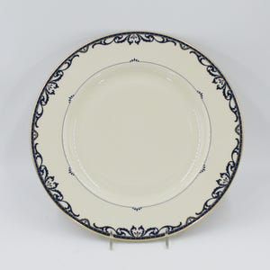 May include: A round, cream-colored plate with a navy blue and gold decorative border. The plate features a delicate, ornate design around the rim and a thin silver line near the center. The plate is displayed on a clear stand.