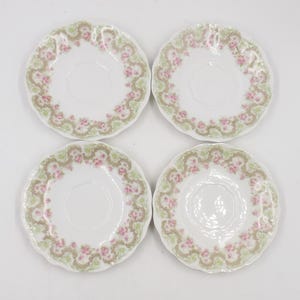 Puede incluir: Cuatro platillos blancos con bordes festoneados. Cada platillo presenta un diseño floral con flores rosas, hojas verdes y un borde marrón. Los platillos están dispuestos en forma cuadrada.