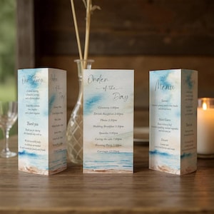 May include: Three-sided wedding stationery with a watercolor beach design. The stationery includes an "Our Story" card, an "Order of the Day" card, and a "Menu" card. The cards are decorated with blue and white watercolor washes.