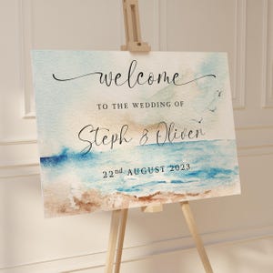 May include: A wedding welcome sign on an easel, painted in watercolour. The sign reads "welcome" in script, followed by "TO THE WEDDING OF Steph & Oliver 22nd AUGUST 2023" over a beach scene with blue water and sand.