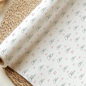 May include: A roll of white wrapping paper with a repeating pattern. The pattern includes the name "Chloe" in a cursive script, the number "21", and small heart shapes in a pinkish-red color. The paper is partially unrolled.