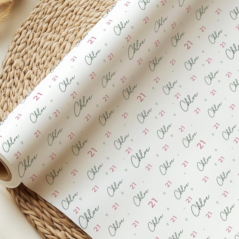 May include: A roll of white wrapping paper with a repeating pattern. The pattern includes the name "Chloe" in a cursive script, the number "21", and small heart shapes in a pinkish-red color. The paper is partially unrolled.