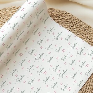 May include: White wrapping paper with a repeating pattern of the name "Sophia" in a script font and the number "21" in a red font. The paper is rolled and placed on a woven surface.
