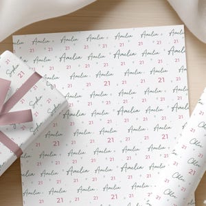 May include: White wrapping paper with a repeating pattern of the name "Amelia" and the number "21" in a handwritten style. A wrapped gift with a pink ribbon is visible on the left.