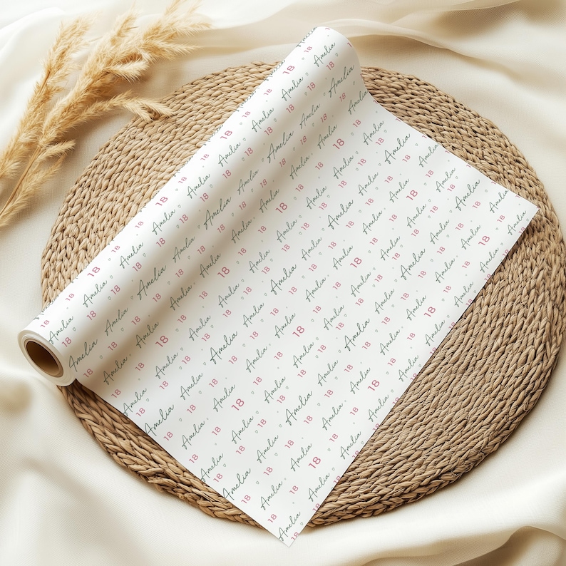 May include: A roll of white wrapping paper featuring a repeating pattern of the name Amelia in a dark script font interspersed with the number 18 in a pink sans-serif font, arranged in diagonal lines across the surface of the paper.