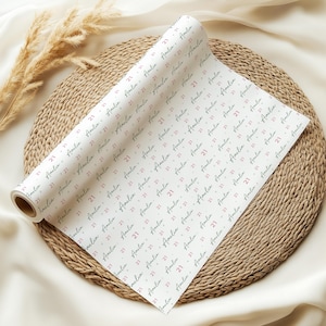 May include: White wrapping paper roll with a repeating pattern of the name "Amelia" and the number "21" in a handwritten style. The paper is on a woven mat with dried plants.