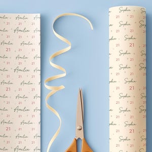 May include: Two rolls of personalized wrapping paper with the names "Amelia" and "Sophia" printed on them, along with the number "21". A cream-colored ribbon and scissors are also on a light blue surface.
