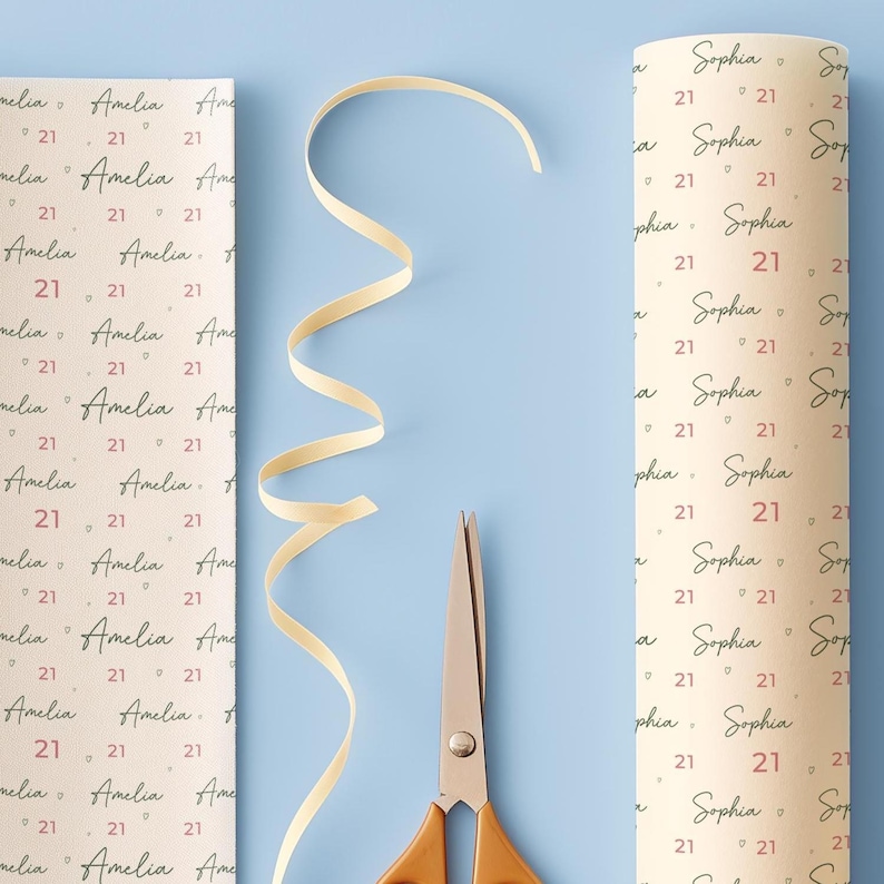 May include: Two rolls of personalized wrapping paper with the names "Amelia" and "Sophia" printed on them, along with the number "21". A cream-colored ribbon and scissors are also on a light blue surface.