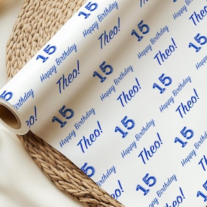 May include: A roll of white wrapping paper featuring a repeating pattern of the number "15", the words "Happy Birthday", and the name "Theo!" in blue. The paper is displayed on a woven, light brown surface.