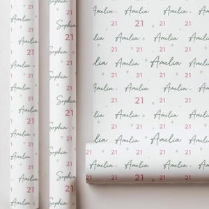 May include: White wrapping paper with a repeating pattern of the name "Amelia" in a handwritten font, the number "21" in a pink hue, and the name "Sophia". The paper is rolled and ready for use.