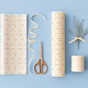 May include: Two rolls of cream-colored wrapping paper featuring repeating black script names and the red number 18, accompanied by a pair of gold-handled scissors, a length of curled cream ribbon, a small spool of twine, and a sprig of greenery.