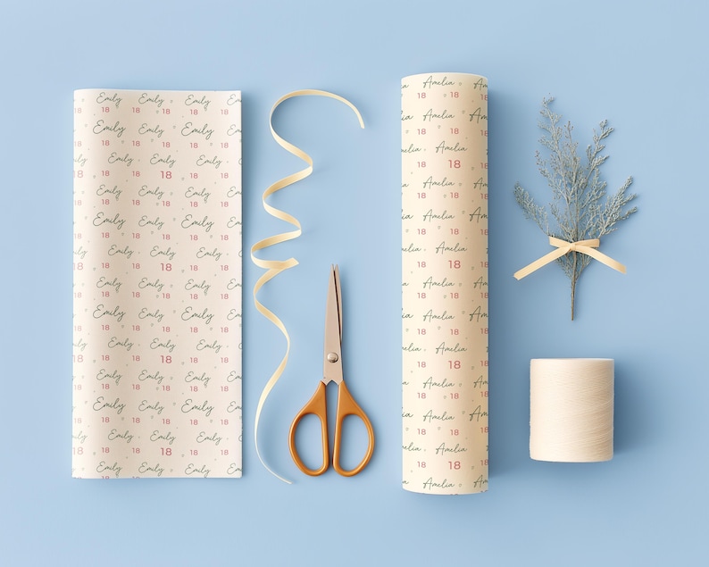 May include: Two rolls of cream-colored wrapping paper featuring repeating black script names and the red number 18, accompanied by a pair of gold-handled scissors, a length of curled cream ribbon, a small spool of twine, and a sprig of greenery.