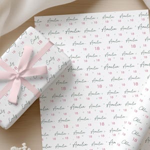 May include: White wrapping paper featuring a repeating pattern of the name Amelia in black cursive script, interspersed with the number 18 in pink, wrapped around a gift box tied with a light pink satin ribbon bow.