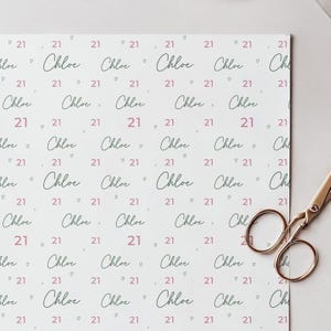 May include: White wrapping paper with a repeating pattern of the name "Chloe", the number "21", and small hearts in pink. A pair of gold scissors is in the bottom right corner. Ideal for a 21st birthday.