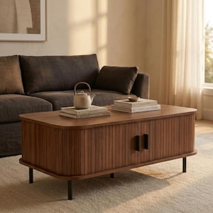 May include: A rectangular wooden coffee table with a slatted front design and two black vertical handles. The table features rounded corners and stands on four thin black legs. Two stacks of books and a small ceramic teapot sit on top.