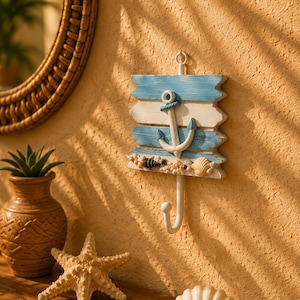 Könnte beinhalten: Ein Wandhaken mit einer rechteckigen Holzplatte, die mit horizontalen blauen und weißen Streifen bemalt ist. Ein weißer Anker ist in der Mitte befestigt, mit einer Ansammlung kleiner Muscheln an der Unterkante und einem einzelnen Metallhaken.