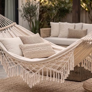 May include: A cream-colored fabric hammock featuring decorative macrame fringe along the edges. The hammock is suspended by thick woven ropes and contains three rectangular throw pillows in shades of cream and beige, providing a soft surface.