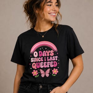 May include: A black short-sleeved t-shirt featuring a graphic print with a pink rainbow, a butterfly, and two flowers. The text reads 0 DAYS SINCE I LAST QUEEFED in pink and white bubble letters, accented by small white stars on the dark fabric.
