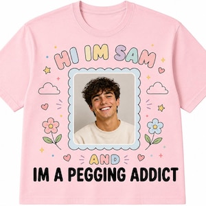 Könnte beinhalten: Ein hellrosa T-Shirt mit einem zentralen rechteckigen Rahmen, der ein Foto einer lächelnden Person enthält. Der Text "HI IM SAM" steht in Pastellfarben darüber, und "AND IM A PEGGING ADDICT" ist in Schwarz darunter gedruckt, umgeben von kleinen Blumen, Sternen und Herzen.