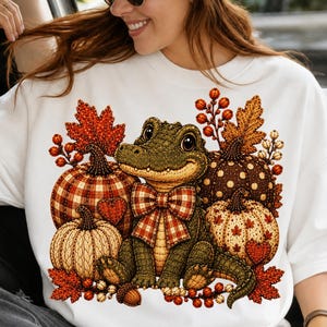 Può includere: Una t-shirt bianca con la grafica di un alligatore verde che indossa un papillon a quadri, circondato da varie zucche con texture a quadri, a pois e lavorate a maglia, insieme a foglie autunnali e bacche nei toni dell'arancione, rosso e marrone.