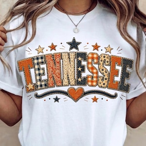 May include: A white t-shirt featuring the word TENNESSEE in large, blocky letters. Each letter has a unique pattern, including plaid, paisley, and cable knit textures in orange, black, and tan. Small stars and a heart accent the design.
